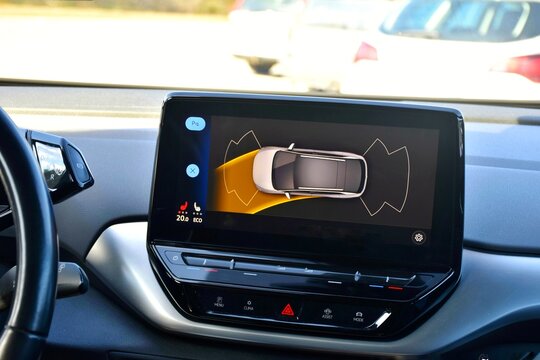The car display screen shows the climate control and parking assistant. A white car is visible, surrounded by parking sensor lines.