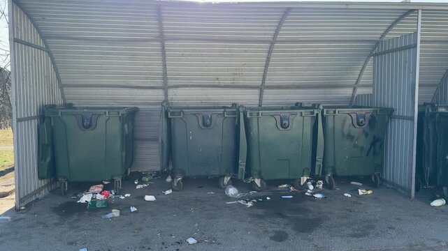 Sunlit covered dumpster area with garbage. Three green bins under metal canopy, litter scattered across stained asphalt, plastic.