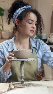 Vertical view of woman placing clay piece on bowl and shaping surface. Female holding small element and adjusting form. Creating handmade pottery and refining details. Crafting process.