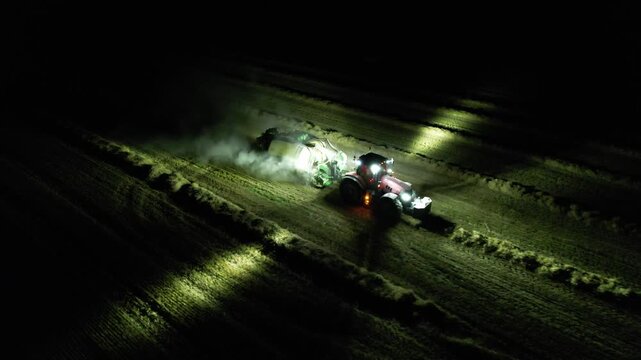Agricultural baler attached to tractor working at night with illuminated lights captured as aerial panorama during hay or straw processing modern technology