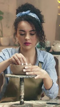 Woman shaping clay bowl with fingers while rotating pottery stand. Female artisan forming ceramic piece and focusing on details. Creating handmade object. Carefully adjusting shape.