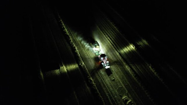 Agricultural baler attached to tractor working at night with illuminated lights captured as aerial panorama during hay or straw processing modern technology