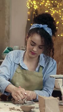 Woman shaping clay with hands while forming ceramic piece on table. Female artisan working with pottery and looking down at material. Creating handmade object. Concentrating on smooth shaping process.