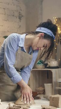Woman leaning over table while shaping clay with hands. Female artisan working on pottery and focusing on process from side view. Creating handmade object in cozy workshop. Vertical view.