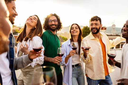 Happy smiling friends enjoying a rooftop party with wine, toasting and laughing together during a warm sunset gathering, sharing moments of joy outdoors