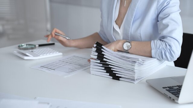 Businesswoman managing heavy workload, organizing documents, accounting for taxes, planning financial reports, processing paperwork, and working with pile of files in modern office. Audit and taxes