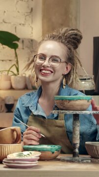 Caucasian woman with glasses smiling and holding ceramic bowl on stand. Female showing finished pottery and enjoying result. Presenting handmade creation and feeling proud. Vertical view.