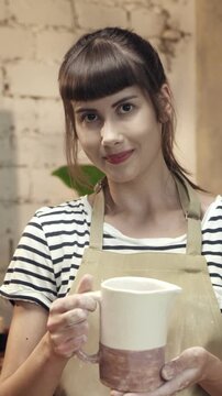 Woman holding ceramic jug while showing handmade pottery to camera. Female artisan presenting crafted object and looking directly forward. Demonstrating finished piece in workshop environment.