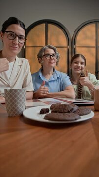 Vertical shot of multigenerational family of three dancing and gesturing on camera while sitting at table with cookies and tea and vlogging at home