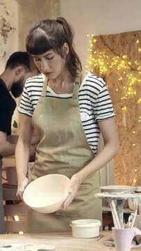 Woman holding ceramic bowl and container while preparing to work on pottery. Female artisan organizing materials. Creating handmade objects. Focusing on preparation and careful process.