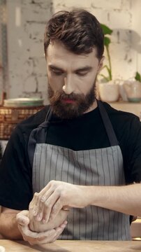 Man shaping clay with hands while focusing on pottery process. Male artisan forming ceramic piece and looking down at work. Concentrating on texture and details of material. Crafting environment.