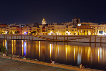 Night view of the Guadalquivir River waterfront with illuminated buildings and reflections in Seville, Spain.
