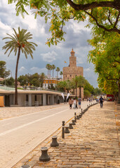 View of Torre del Oro along the riverside promenade with palm trees, pedestrians and dramatic cloudy sky in Seville, Spain.