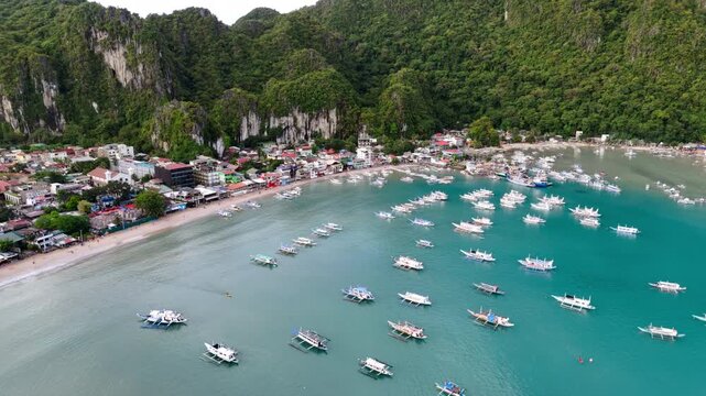 Crane tilt-down drone shot of tourist boats lined up at El Nido Bay in the morning, revealing calm turquoise waters, limestone mountains, and a small coastal town, Philippines