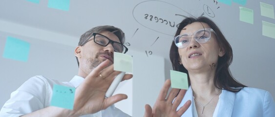 Business colleagues collaborating on strategy, brainstorming ideas for success, writing on a transparent glass board with sticky notes in a modern office © volha_r