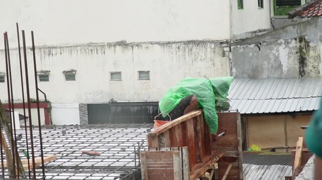 Construction worker in green raincoat working on steel reinforcement during rain. Industrial labor and building site development under difficult weather conditions concept. asian worker