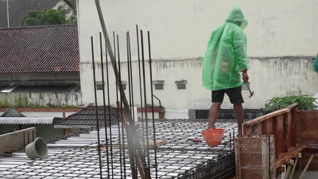 Construction worker in green raincoat working on steel reinforcement during rain. Industrial labor and building site development under difficult weather conditions concept. asian worker