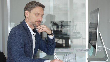 Fototapeta premium Businessman in a blue jacket working on computer, typing and writing notes in modern office, managing tasks, multitasking. Business people concept