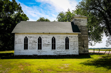 Obraz premium An old white wooden church stands on green grass surrounded by trees with a bright blue sky weathered walls and tall windows create a peaceful rural atmosphere
