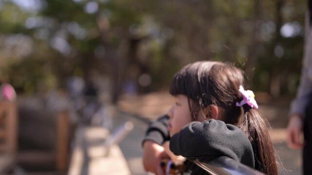 Child gazes thoughtfully at distant scenery. Hair tied back with a soft purple flower. Child leans against a metal railing. Bright sunlight filters through trees