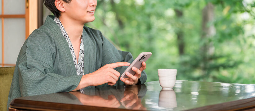 A man in a yukata (traditional Japanese robe) is looking at his smartphone in a Japanese-style room at a ryokan (traditional Japanese inn).
