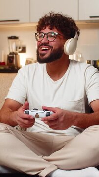 Arabian man at home plays on game console holds joystick leans forward enjoys play sees level score high. Young guy on sofa cheers after win presses buttons shoots jumps feels thrill night victory.