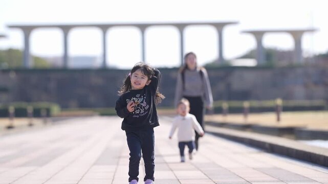 Sunlight glows softly over a paved path. Young girl dances joyfully, arms wide. Her mother and baby follow, blurred in the background. The arches stretch into the gentle distance