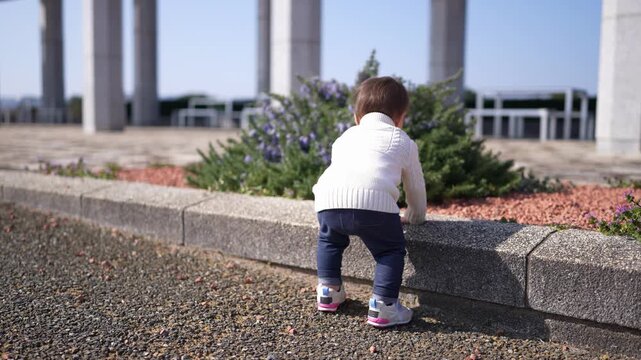 Little child explores sidewalk curbs, curious and independent. Bright sunlight illuminates the scene, casting gentle shadows. Concrete pillars frame a modern, open space behind the child