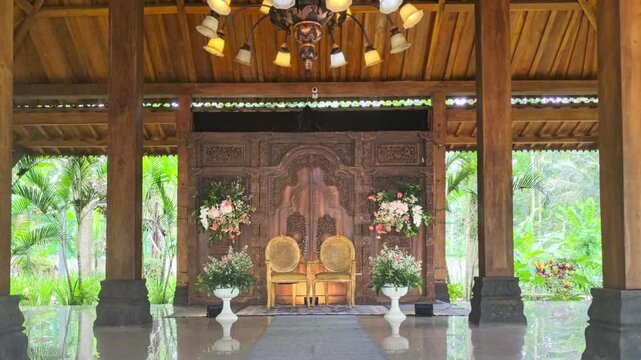 Traditional Javanese Wedding Stage in Indonesia with Gebyok Carved Wood Background, Elegant Chairs, Floral Decoration, and Luxury Chandelier in Open Pavilion Joglo House Java