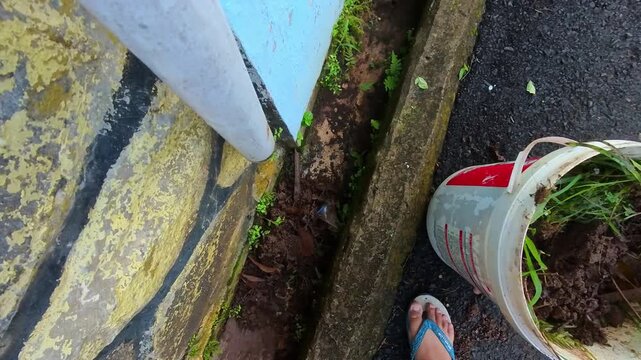 Overhead clip of a man digging a drain with a hoe and pouring the soil into a bucket. Video of a man cleaning a house gutter of weeds and dirt. First-person POV footage with steady 4K resolution.