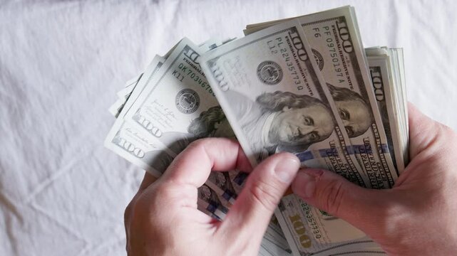 Person's hands counting a large stack of paper currency on white textured fabric. Scene conveys feelings of wealth and abundance during financial transaction.