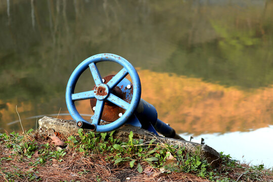 A close-up shot of a rustic, weathered blue industrial valve handle with significant rust and peeling paint, situated on a concrete structure at the edge of a calm body of water. The foreground is ado
