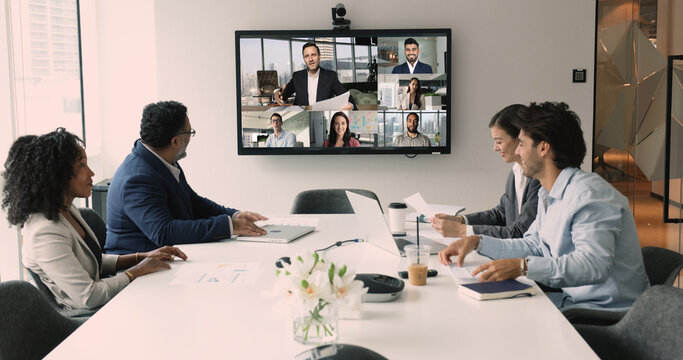 Diverse international business team discussing teamwork, work strategy, planning deal, partnership on online hybrid video conference, meeting at table and large screen, talking