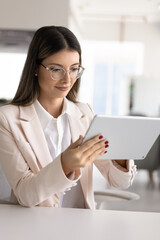 Confident businesswoman in suit using modern digital tablet device sits at desk in modern office,...