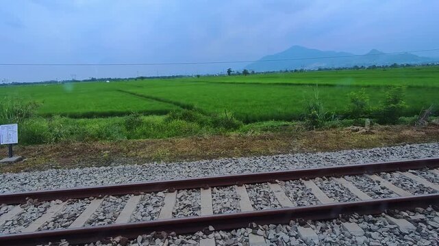 Slow-motion clip showing a train track, consisting of iron rails with concrete sleepers and a gravel stone base, amidst green rice paddies. Steady slow motion in 4K.