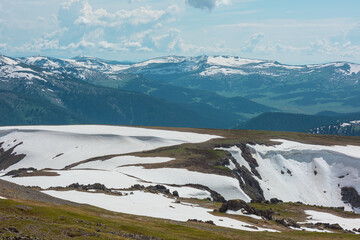 Scenic alpine landscape with sunlit snowy field on stony hill with view to big mountain range with forest and snow far away under cloudy sky. Stone outcrops among snows in sunlight in high mountains. © Daniil