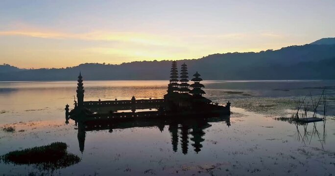 A breathtaking aerial shot of Pura Ulun Danu Tamblingan temple in Bali at sunrise. The spiritual architecture is silhouetted against the colorful sky, with a perfect reflection on the calm lake.