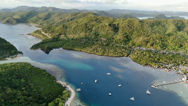 Morning panning aerial shot above Linapacan Island revealing blue waters, a coastal community, and mountainous shoreline terrain at sunrise, Philippines