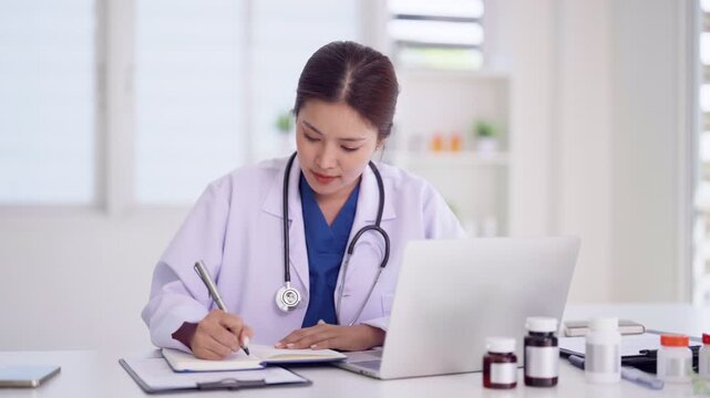 Asian adult woman doctor medical professional focused work laptop writing notes healthcare office environment
