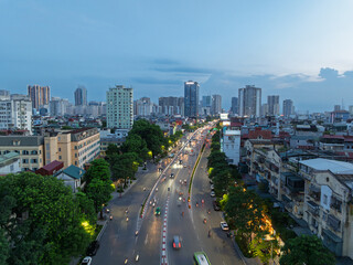 Naklejka premium Aerial evening view of Tran Duy Hung street in Hanoi showing busy traffic flow, tree lined avenue, dense urban buildings, and illuminated cityscape under blue hour sky.