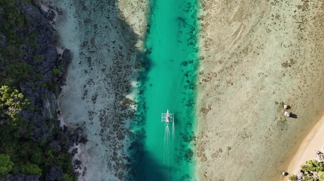 Overhead tracking drone shot of an outrigger boat navigating a narrow crystal-clear channel between islands in Linapacan, Palawan, during morning calm, Philippines