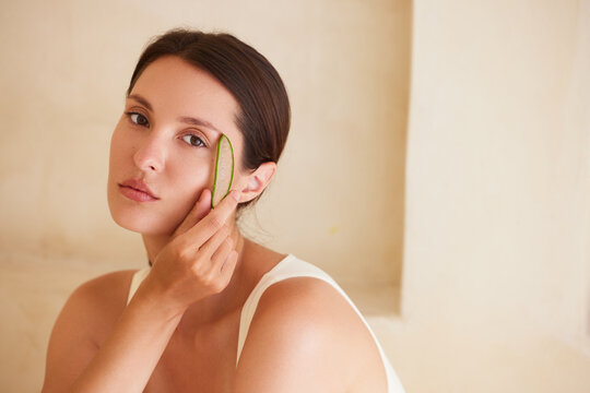 Calm young brunette woman applying a fresh cut aloe vera leaf to her cheek with a serene sideways gaze. Natural botanical skincare concept, beige architectural background, large copy space right. 