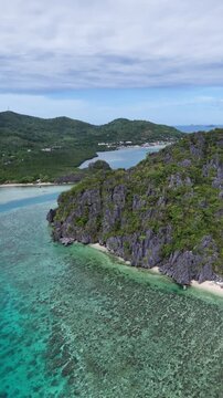 Portrait orientation pull-away panning drone shot over Ile Island showing limestone formations, turquoise waters below, and Linapacan town in the distance, Philippines