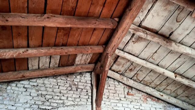 Weathered white brick wall with cracks beneath aged wooden beams and planks forming a triangular roof structure, illuminated by daylight, showing rustic building detail