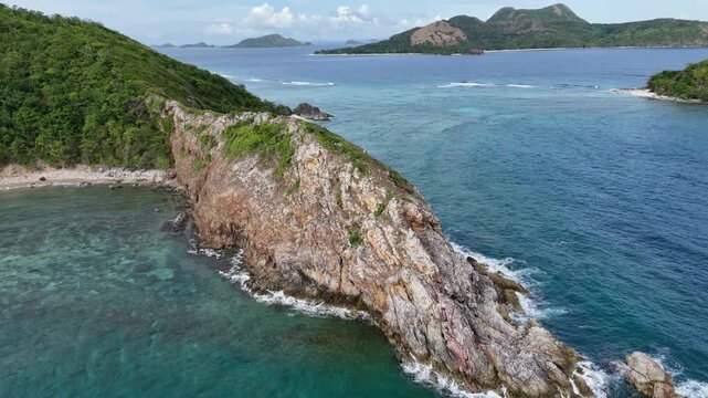 Pull-away drone shot of a scenic cape at Ile Island featuring Kinulay and Patoyo Islands against a vibrant blue ocean and sky, Philippines