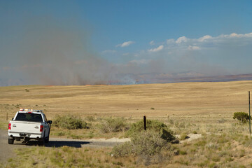 White emergency response pickup truck with flashing lights monitoring a distant wildfire in a dry rural grassland landscape © Scott