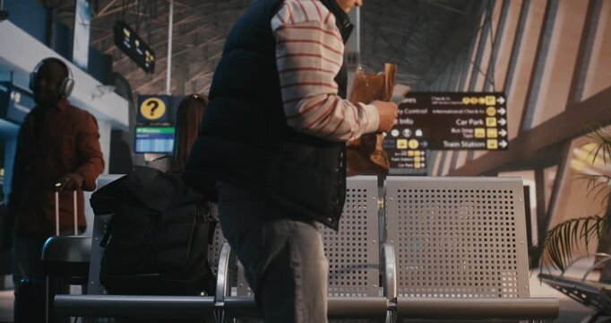Young Man in Striped Shirt And Navy Vest Sits in Airport Terminal, Enjoying Pastry From Paper Bag. With Backpack on Neighboring Seat, Male Looks Around Slowly While Eating. Relatable Travel Moment.