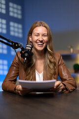 Portrait of smiling female host sitting at studio desk, holding script or notes papers while...