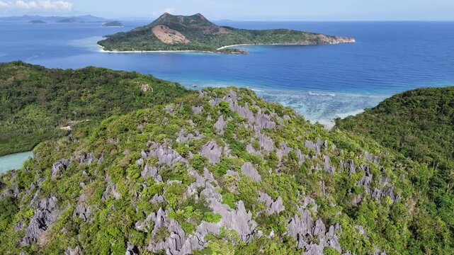 Flyover drone shot of limestone cliffs covered with lush vegetation at Ile Island, Linapacan, revealing a scenic view, a clear shallow channel, and Patoyo Island ahead, Philippines