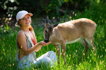 Obraz premium Girl sitting in grass feeding deer. Child connecting with animal in natural environment. Peaceful scene with wildlife interaction. Summer outdoor lifestyle with harmony concept.